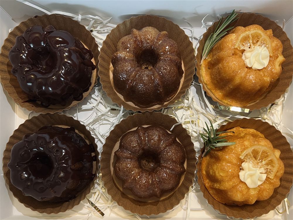 Overhead view of a box of six mini bundt cakes in kraft paper liners: Chocolate Fudge, Apple Dapple and a citrus bundt with rosemary